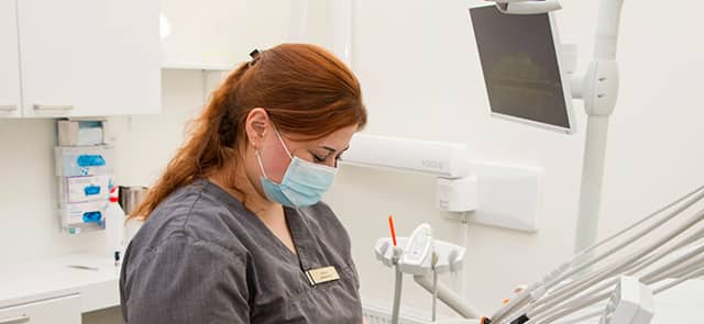 Tandvårdspersonal med munskydd och arbetskläder arbetar i ett behandlingsrum, med dental utrustning och en skärm i bakgrunden.